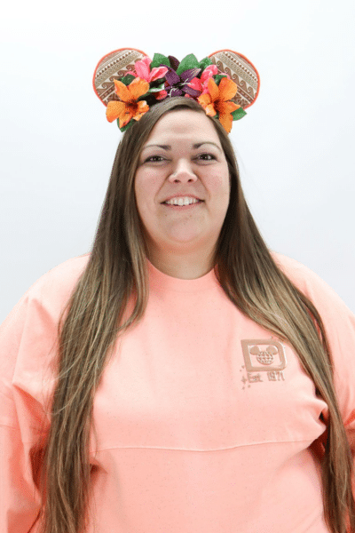 Woman wearing pink shirt and colorful floral ears headband, smiling at camera in studio setting.
