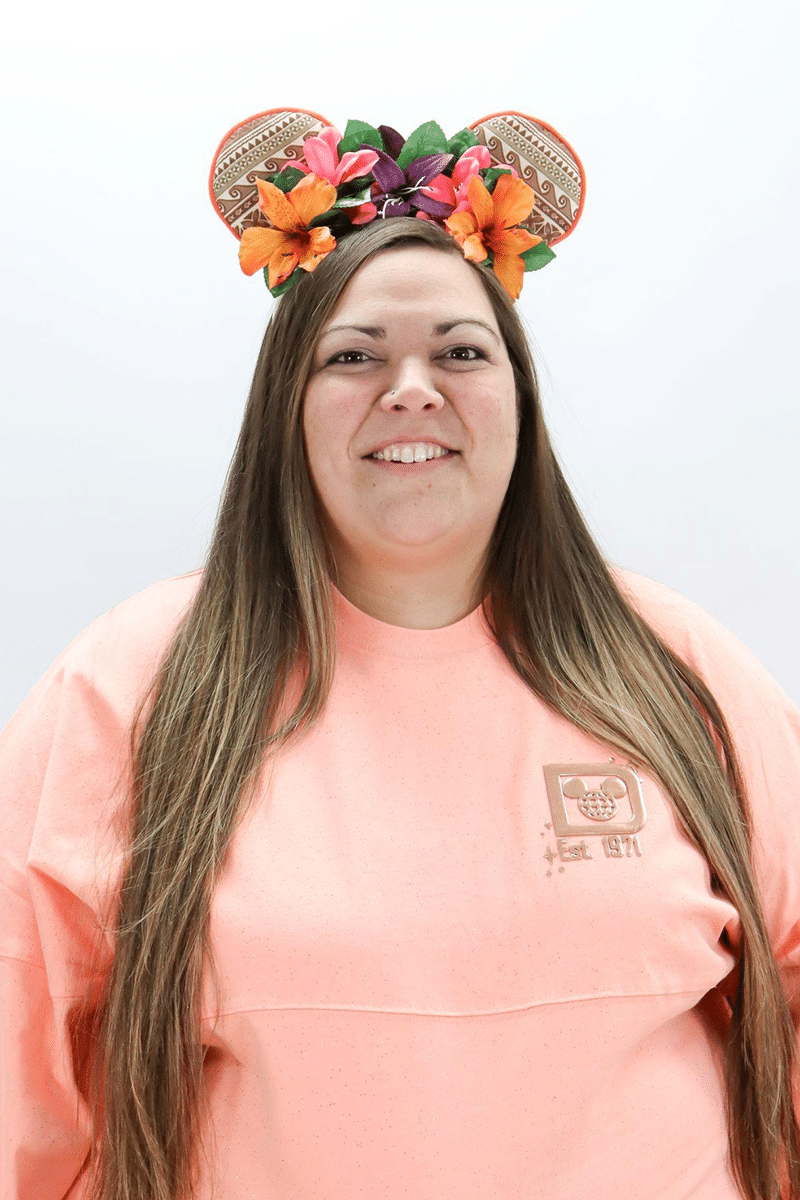 Woman wearing pink shirt and colorful floral ears headband, smiling at camera in studio setting.