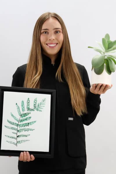 Woman in black blazer holding framed fern print and potted plant against white background.