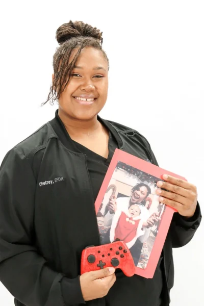 Chelzey, FDA team member at Peachtree City dental practice, smiling while holding family photo and red gaming controller.