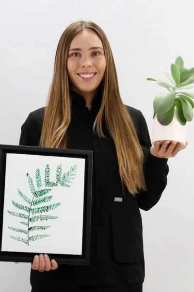 Young female dentist smiling while holding a potted succulent plant and framed botanical fern artwork.