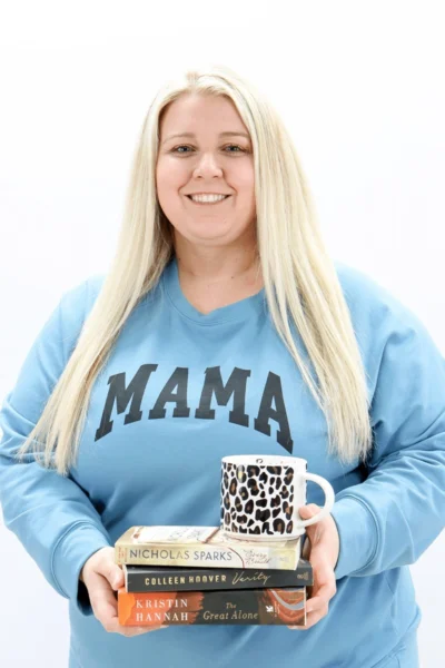 Woman in blue mama sweatshirt holding stack of books and leopard print mug, smiling at camera.